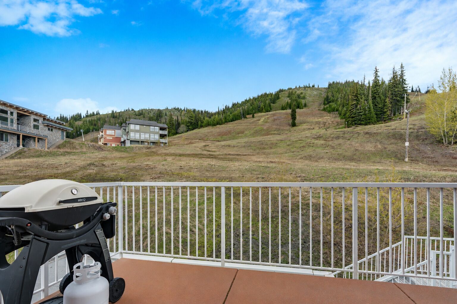 Ein Balkon mit Grill, Gasflasche und Blick auf eine hügelige Landschaft mit Gebäuden.