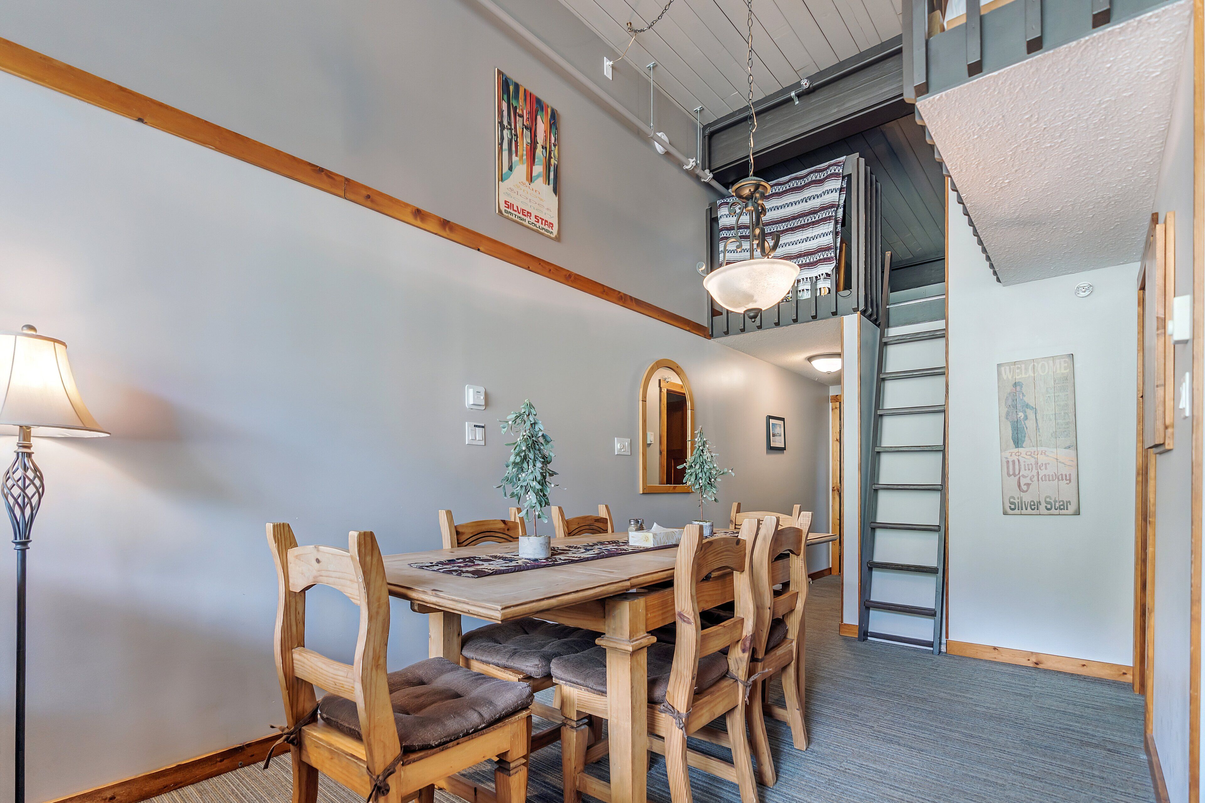 Un espace repas avec une table et des chaises en bois, une lampe et une échelle menant à une mezzanine.