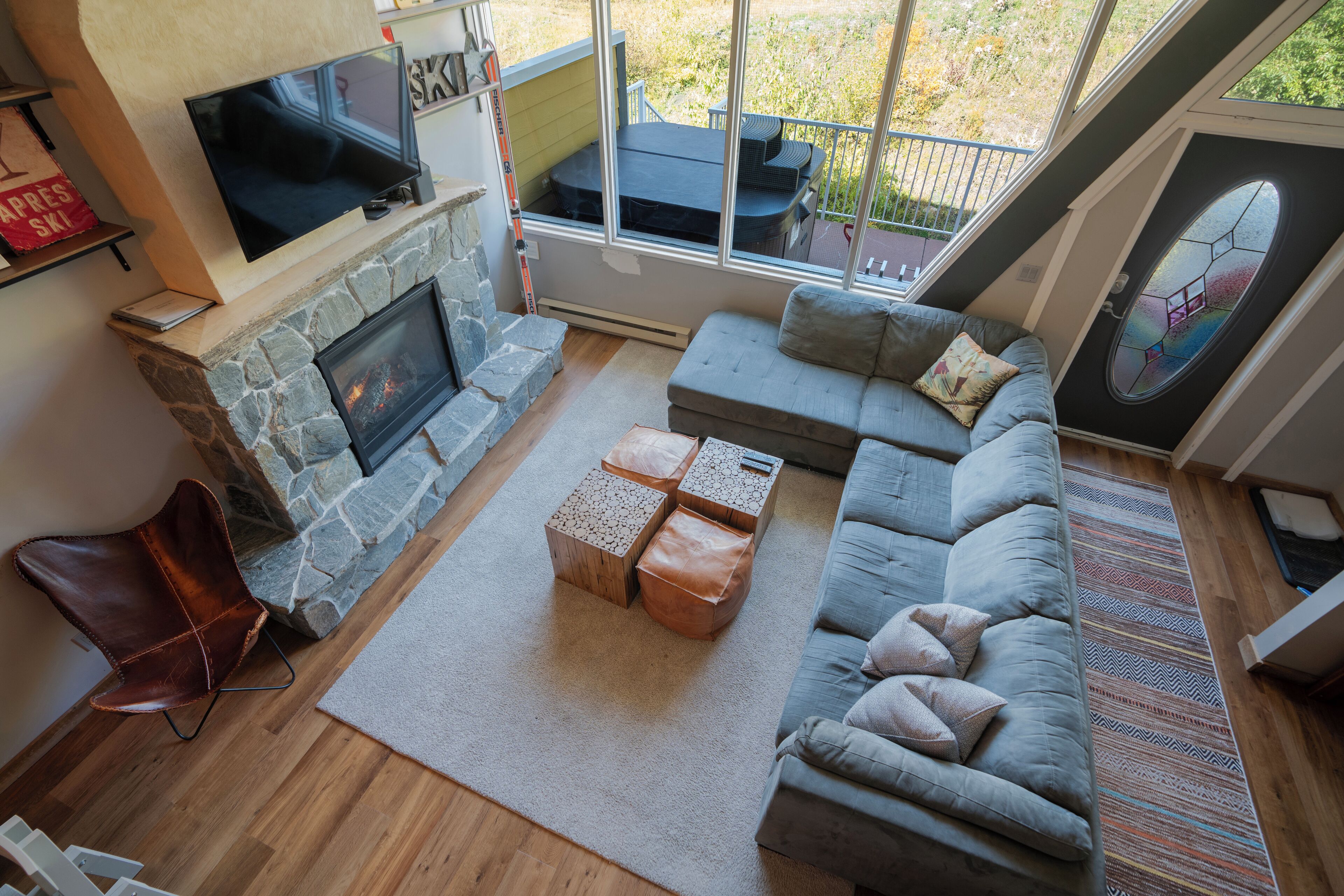 A living room with a stone fireplace, a grey sofa, a TV mounted above the fireplace, and a large window overlooking a deck.