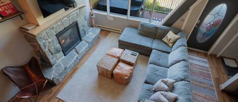 A living room with a stone fireplace, a grey sofa, a TV mounted above the fireplace, and a large window overlooking a deck.