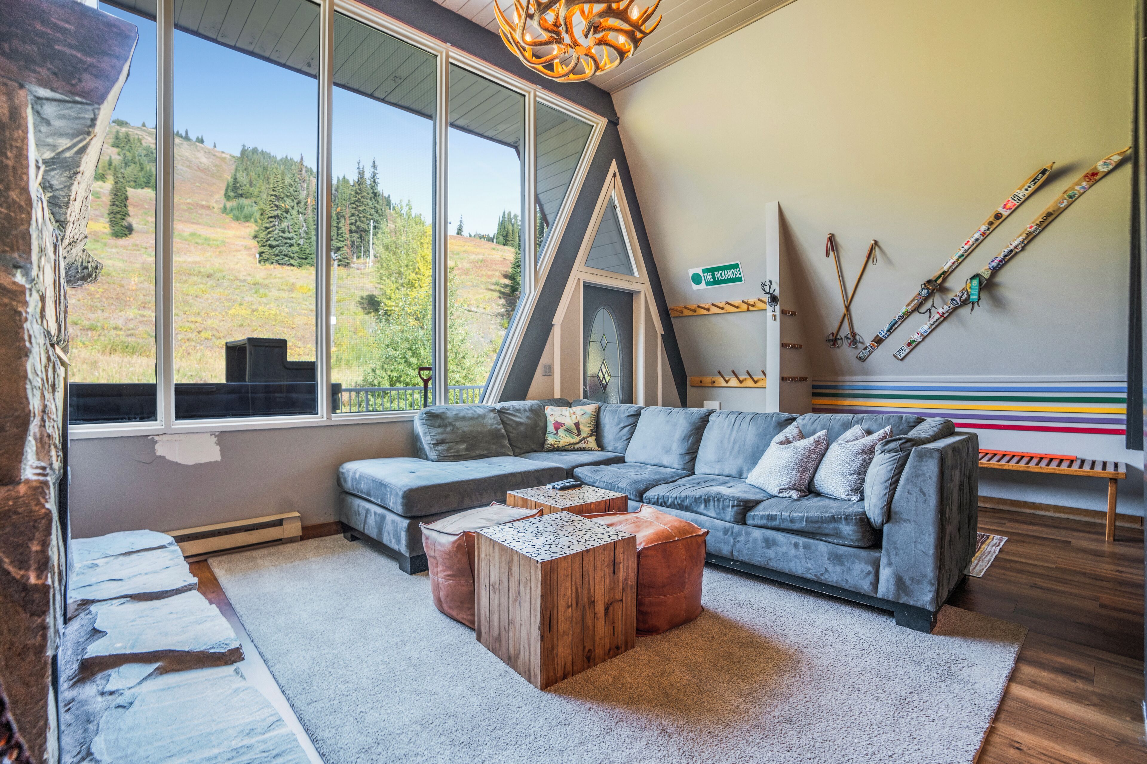 A modern living room with a large window, a sectional sofa, a wooden coffee table, and a ski rack against the wall.