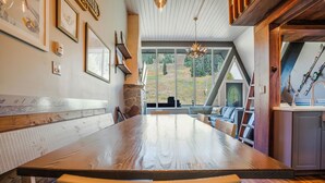 Une table à manger en bois, entourée de chaises, une cheminée en pierre et une vue sur l’extérieur grâce à de grandes fenêtres.
