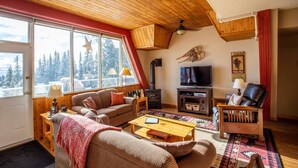 A cozy living room with a wooden ceiling, a fireplace, a television, and a large window overlooking trees.