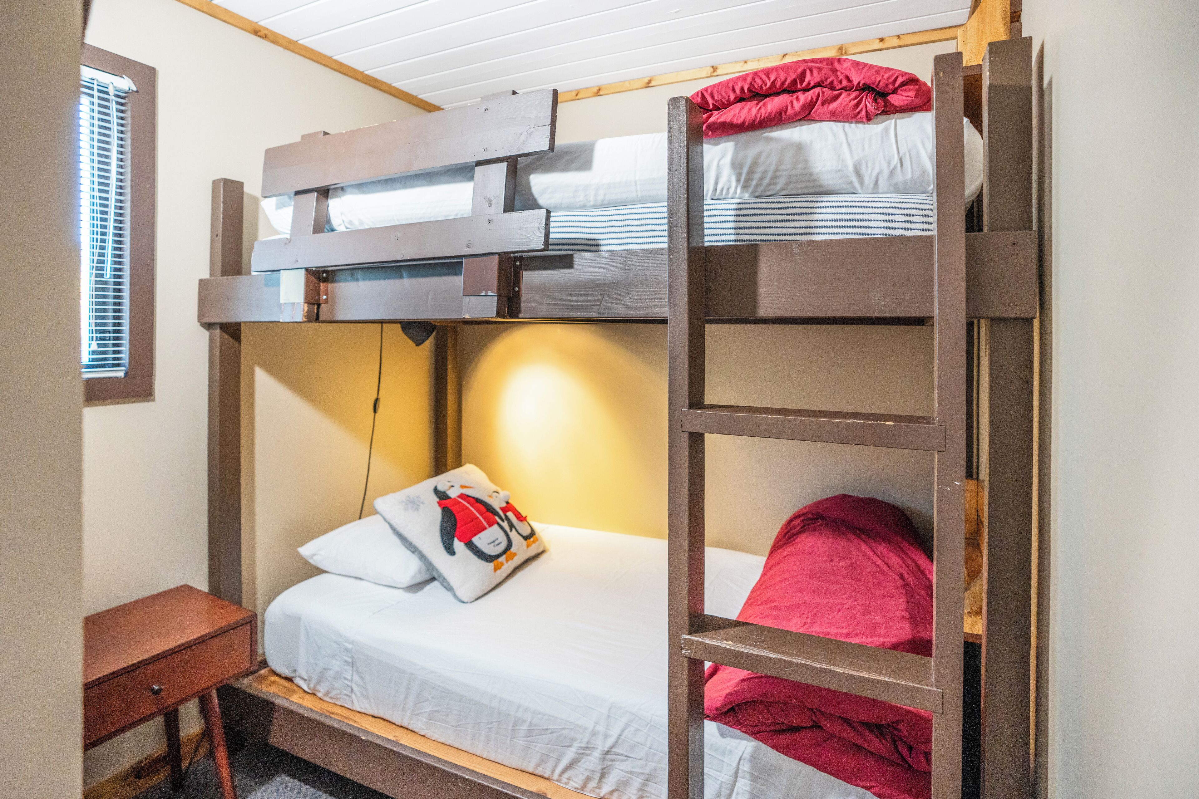 Een slaapkamer met een stapelbed, een klein houten nachtkastje en een raam met jaloezieën.