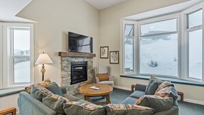 A cozy living room with a stone fireplace, a television mounted above it, and large windows offering a view of a snowy landscape.