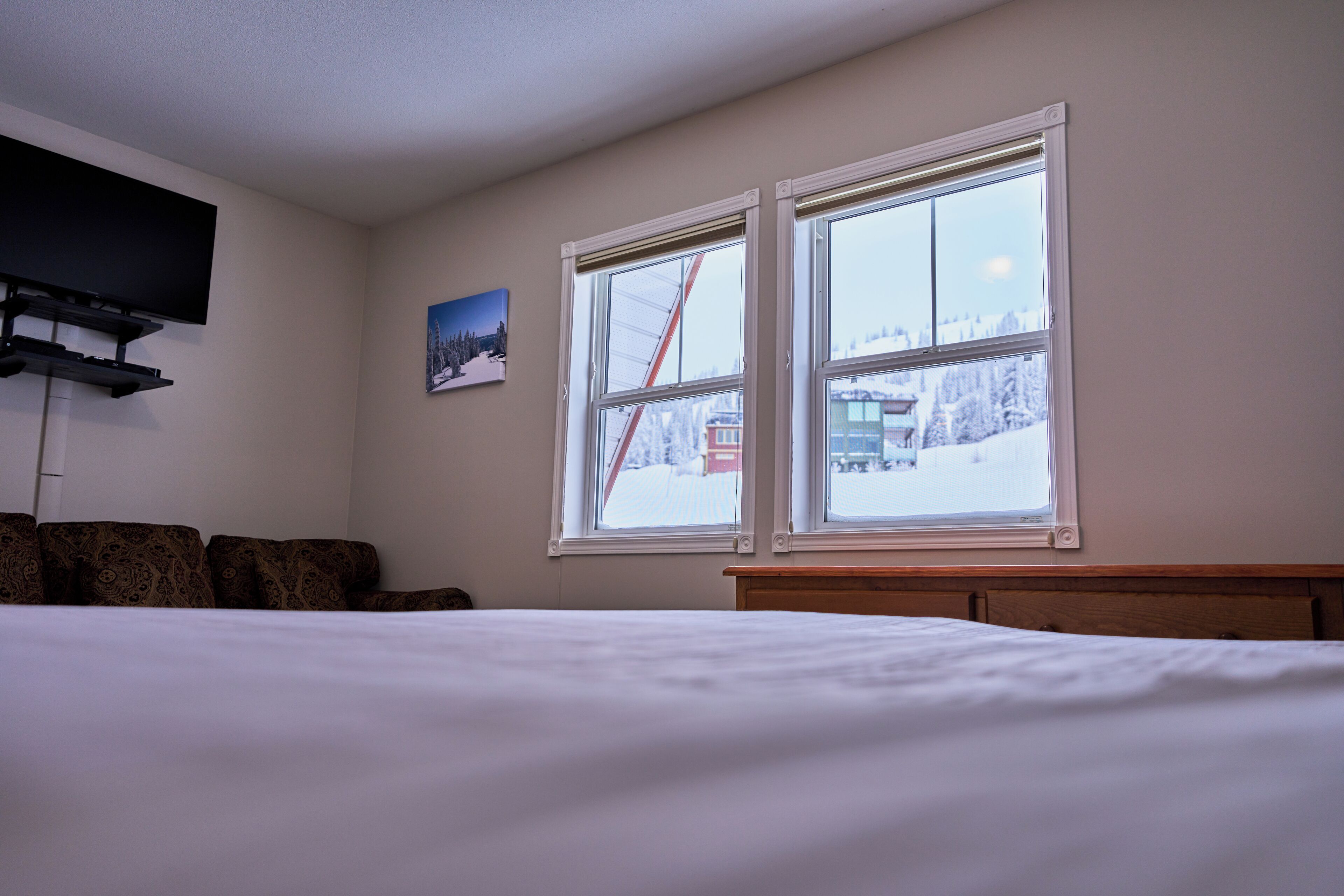 Un dormitorio con cama, televisor montado en la pared y dos ventanas con vista nevada.