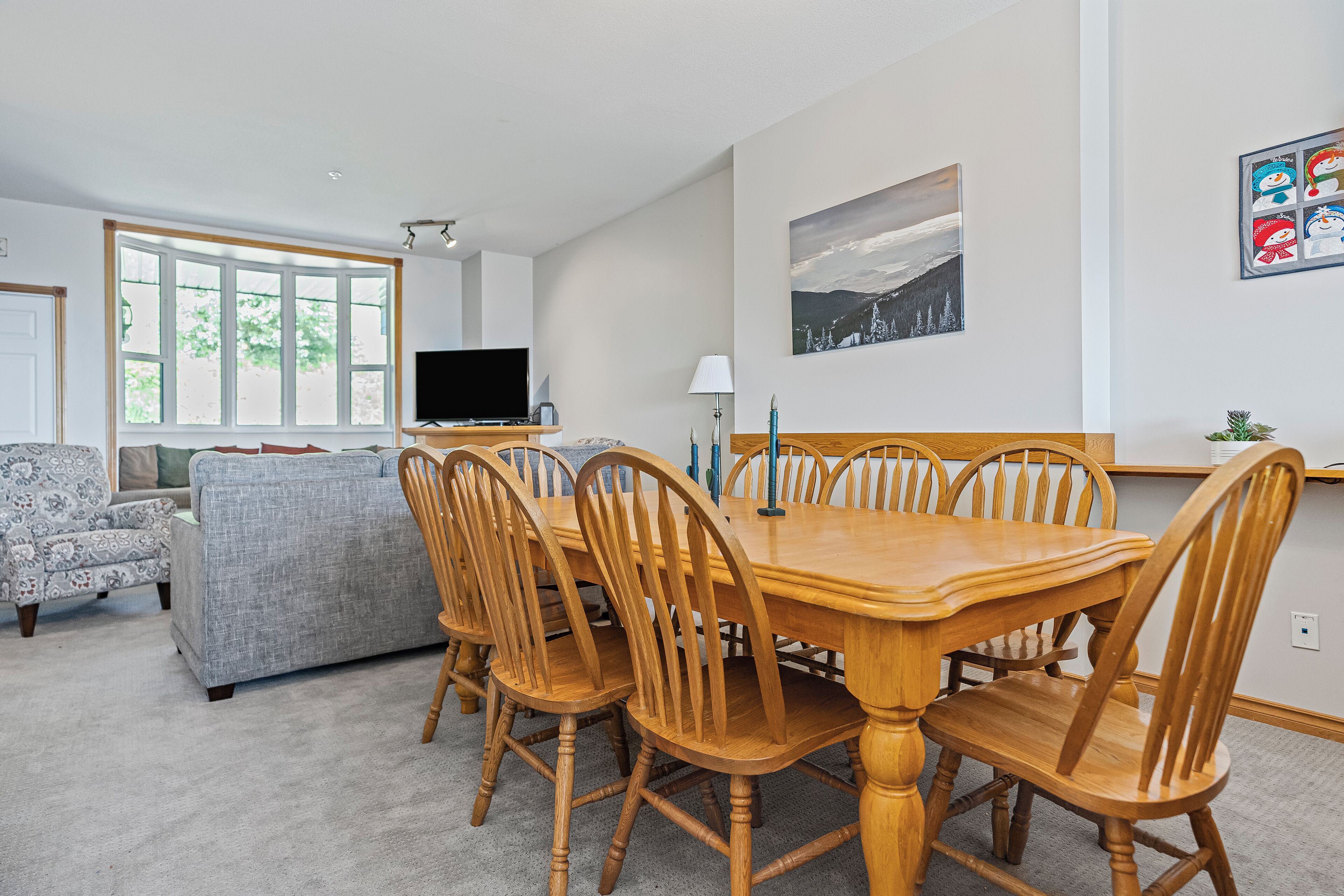 Un comedor con mesa y sillas de madera, televisor y sofá en una habitación luminosa con ventanas amplias.