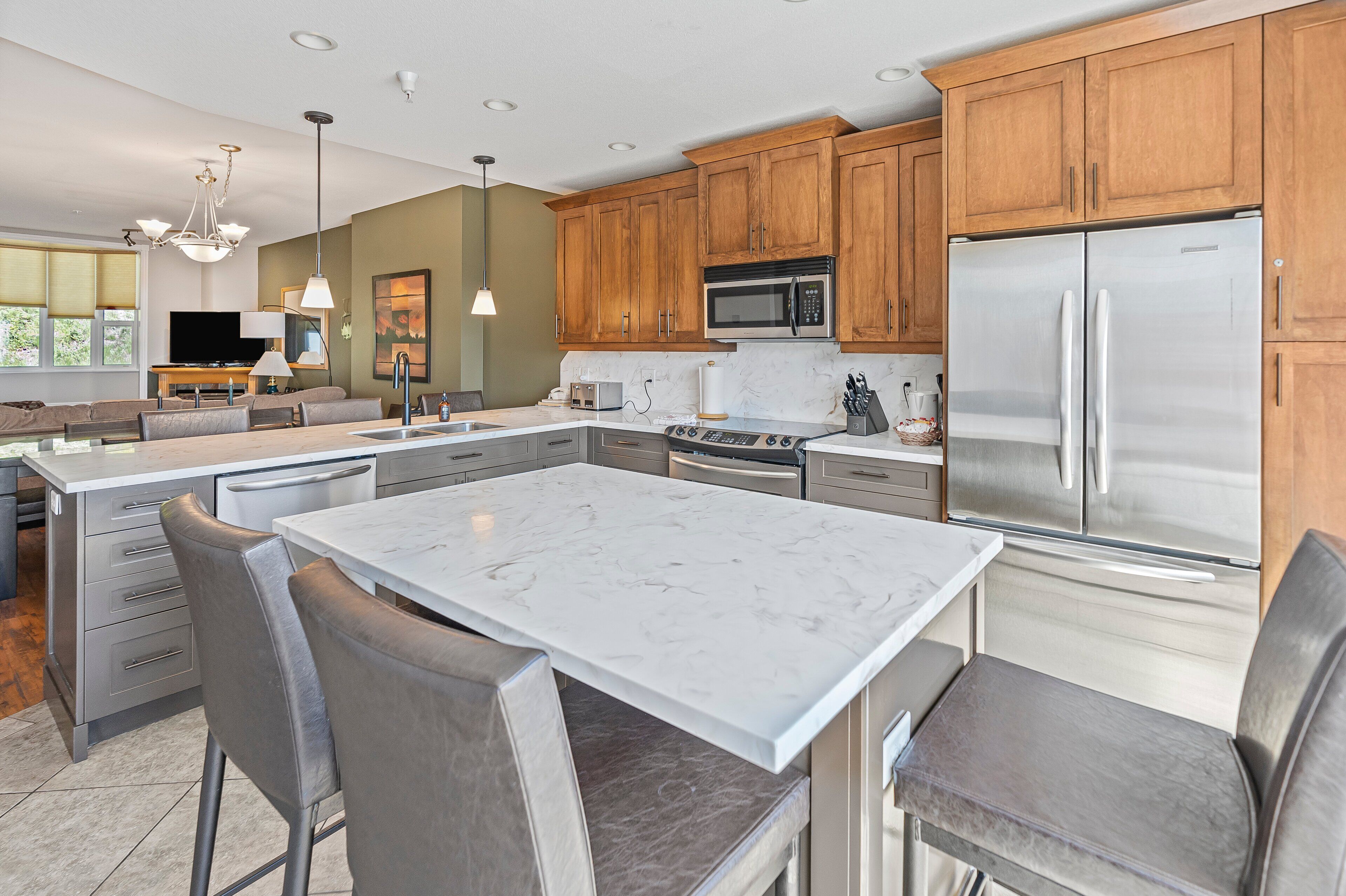 A modern kitchen with wooden cabinets, stainless steel appliances, a marble island, and a dining area with gray chairs.