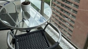 Un balcon avec une table et des chaises en verre, une plante en pot et une vue sur un paysage urbain avec des bâtiments et de la verdure.