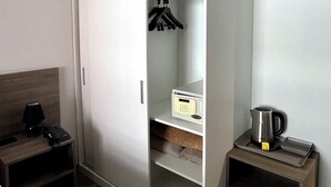 Une chambre d’hôtel avec une armoire blanche, une table de chevet avec une lampe, une petite étagère avec une bouilloire et une tête de lit en bois.