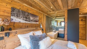 Une chambre à coucher de style cabane en bois, avec un lit, des oreillers, un plaid moelleux, un tableau représentant un paysage enneigé, et une salle de bain avec une baignoire et un lavabo.