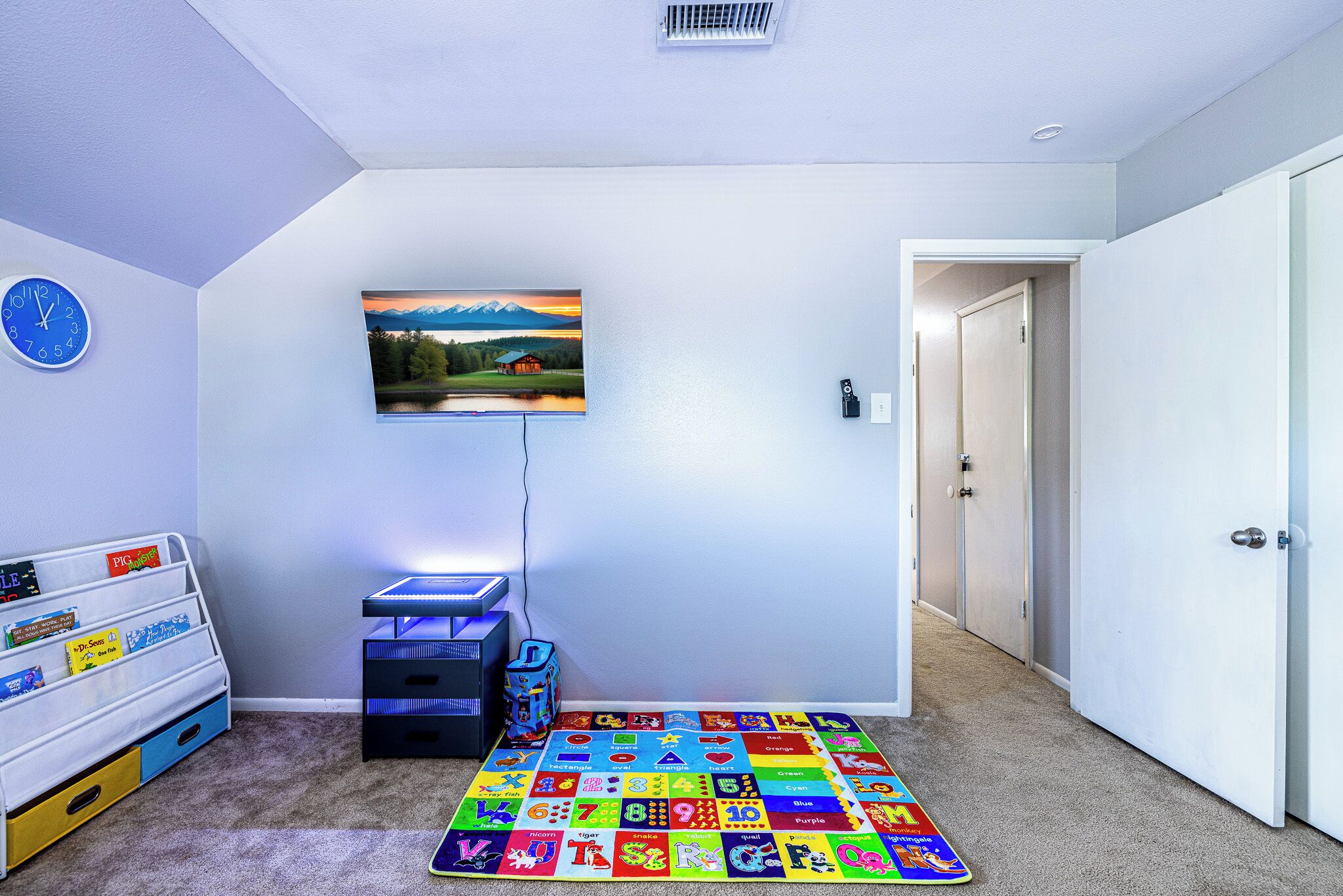 Une salle de jeux pour enfants avec un tapis coloré représentant l’alphabet, une horloge et un tableau représentant un paysage de montagne.
