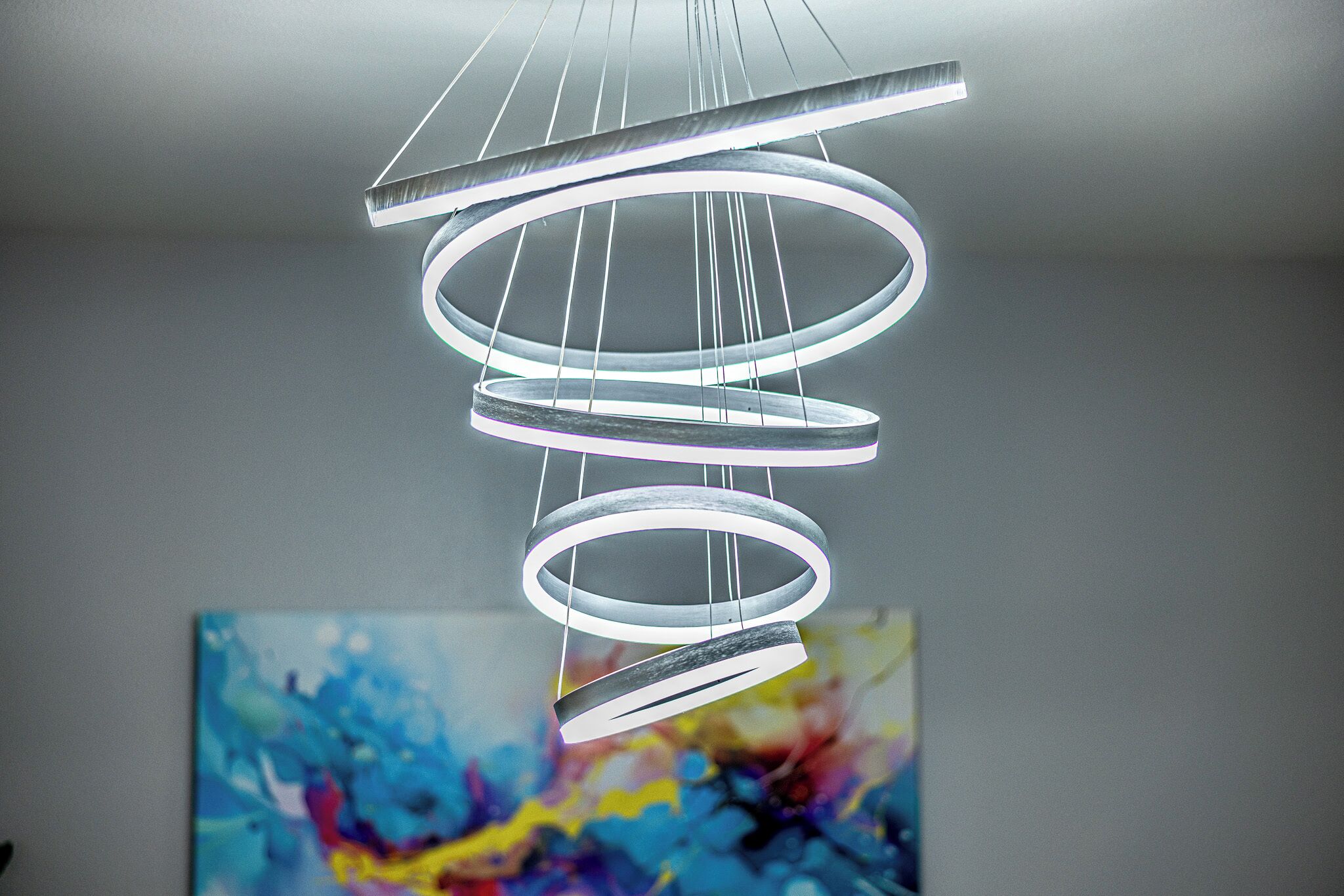Un lustre moderne composé de plusieurs anneaux et un tableau aux couleurs abstraites, dans les tons de bleu et de jaune, accroché au mur.