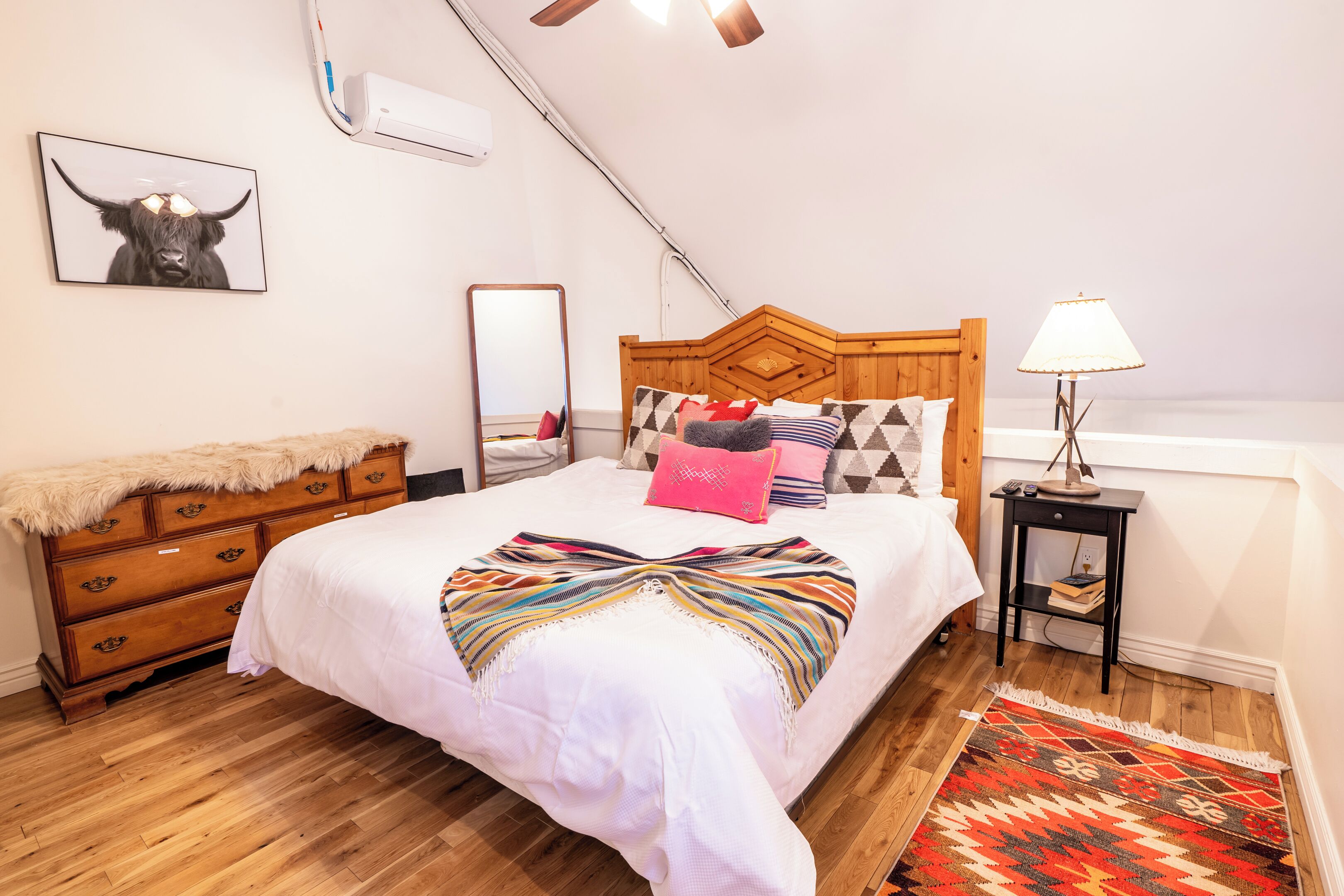 Un dormitorio con cabecera de madera, cama con cubrecama blanco y almohadas coloridas, cómoda de madera con una manta de piel, un espejo, una lámpara en una mesita de noche y una alfombra estampada.