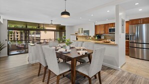 A modern kitchen with stainless steel appliances, a dining area with a wooden table and grey chairs, and a view of a balcony with plants. - Luxurious and Spacious 2BD Kaanapali Condo (Lahaina)