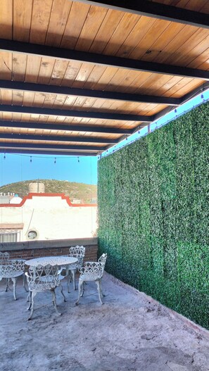 Een terras buiten, met een houten plafond, een groene haag als wand en een tafel met vier stoelen.