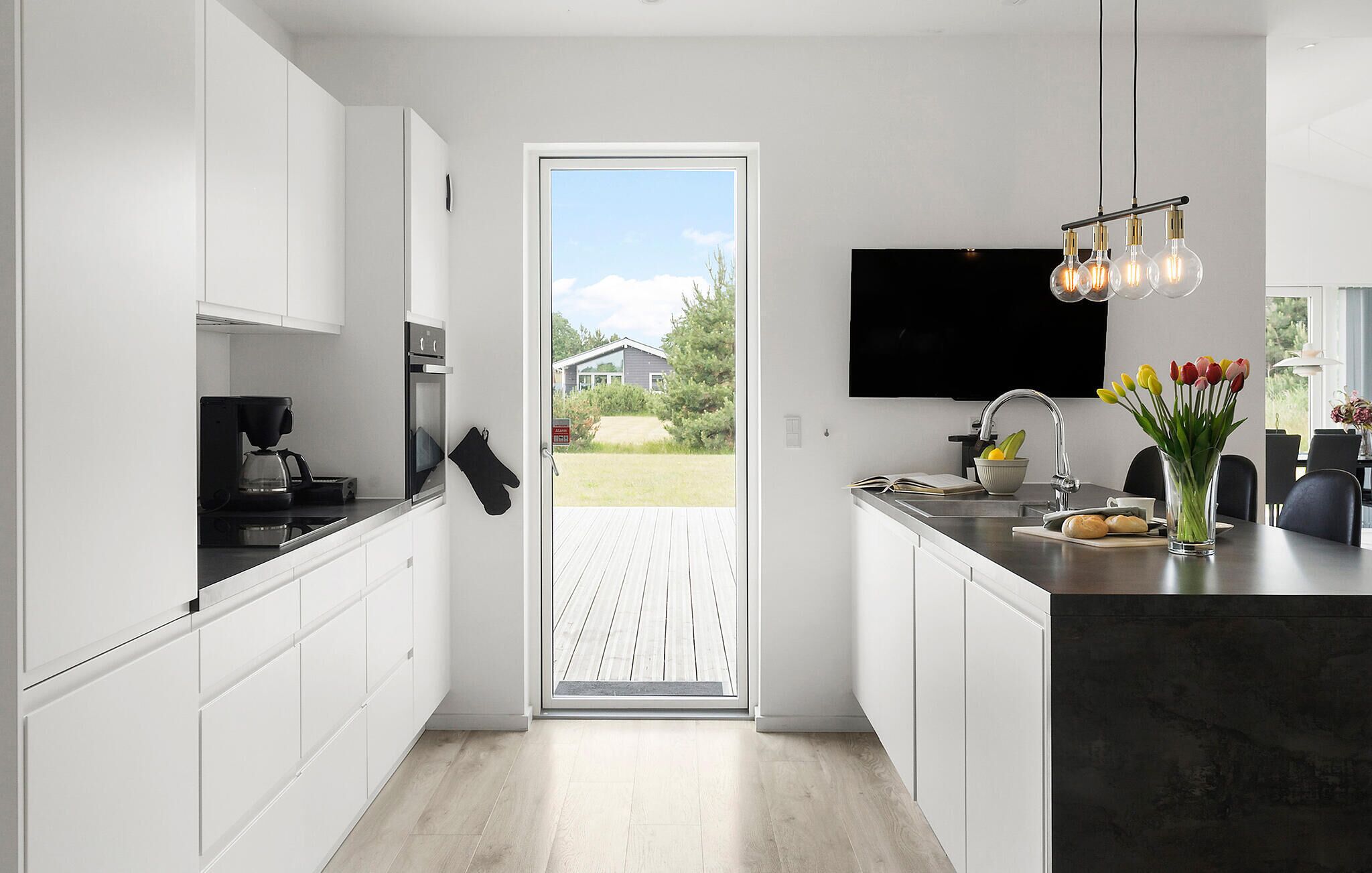 Une cuisine moderne avec des meubles blancs, un four encastré, une machine à café et une porte vitrée donnant sur une terrasse extérieure.