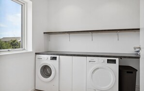 A laundry room with two white front-loading washing machines, a small trash bin, and a window providing a view of the outside.