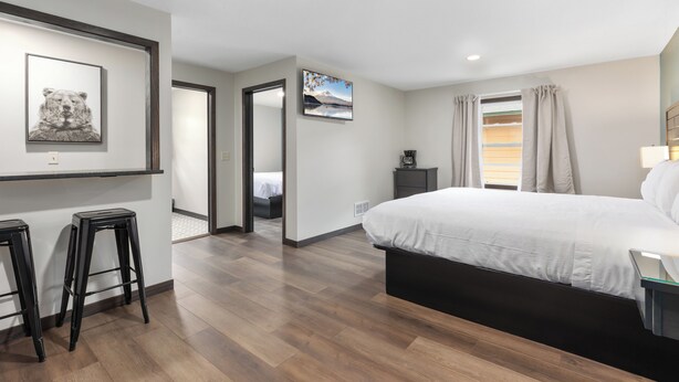 A modern bedroom with a large bed, wooden flooring, a desk with a chair, and a small stool.