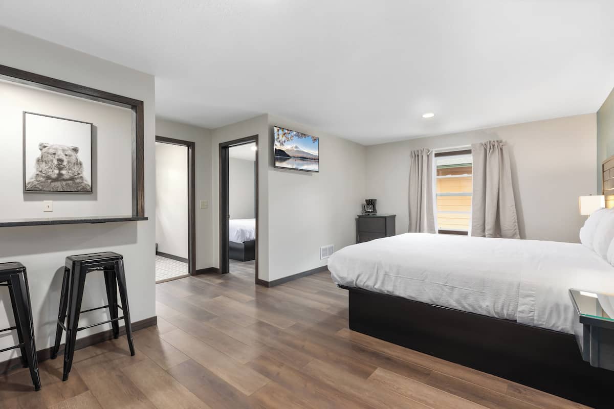 A modern bedroom with a large bed, wooden flooring, a desk with a chair, and a small stool.
