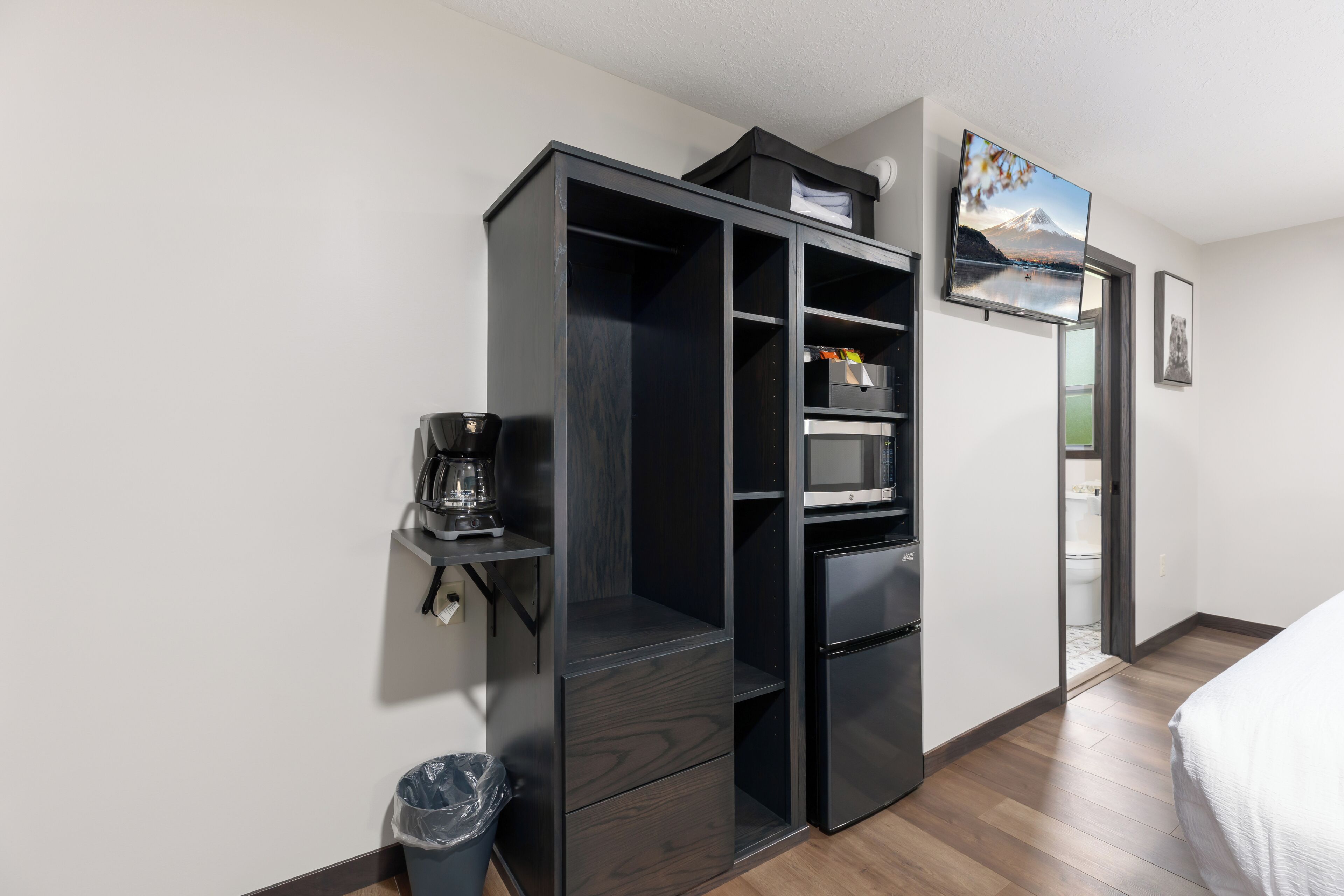 A compact room with a kitchenette, a bed, a trash bin, and a door leading to a bathroom.