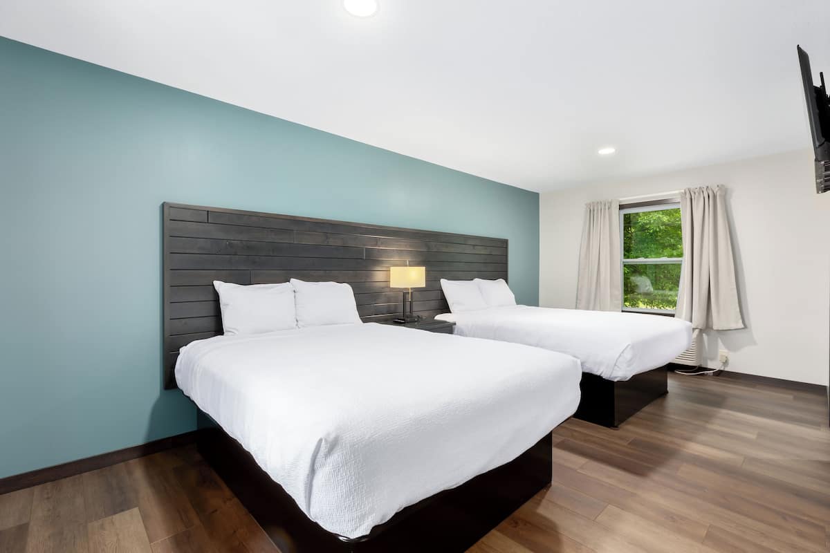 A modern hotel room with two beds, a wooden headboard, a nightstand with a lamp, a window with curtains, and a view of greenery outside.