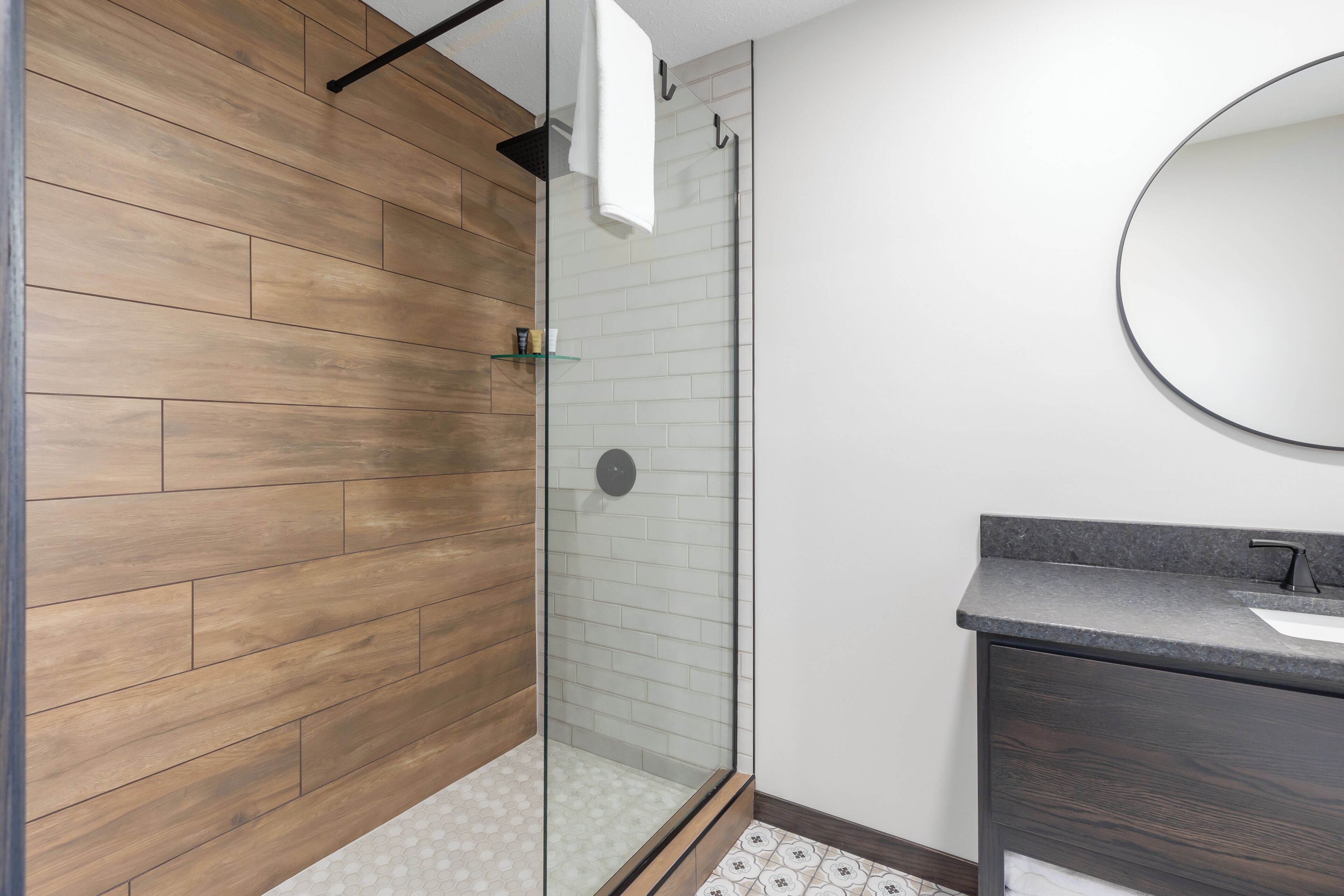 Un baño moderno con pared de madera, cerramiento de ducha de vidrio y tocador de tonos oscuros con un espejo redondo.
