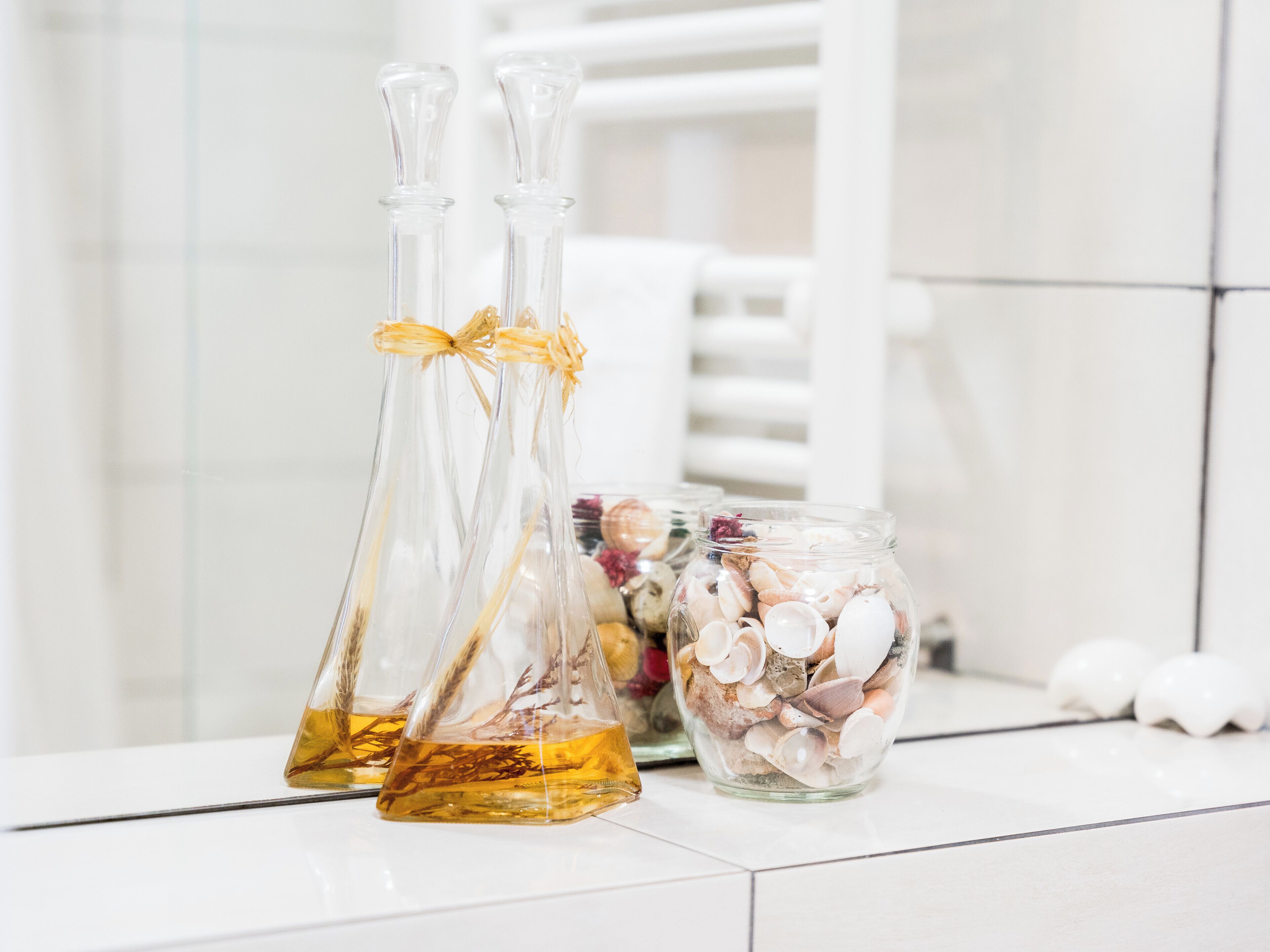 Two glass bottles with amber liquid and a glass jar filled with seashells on a white tiled surface.