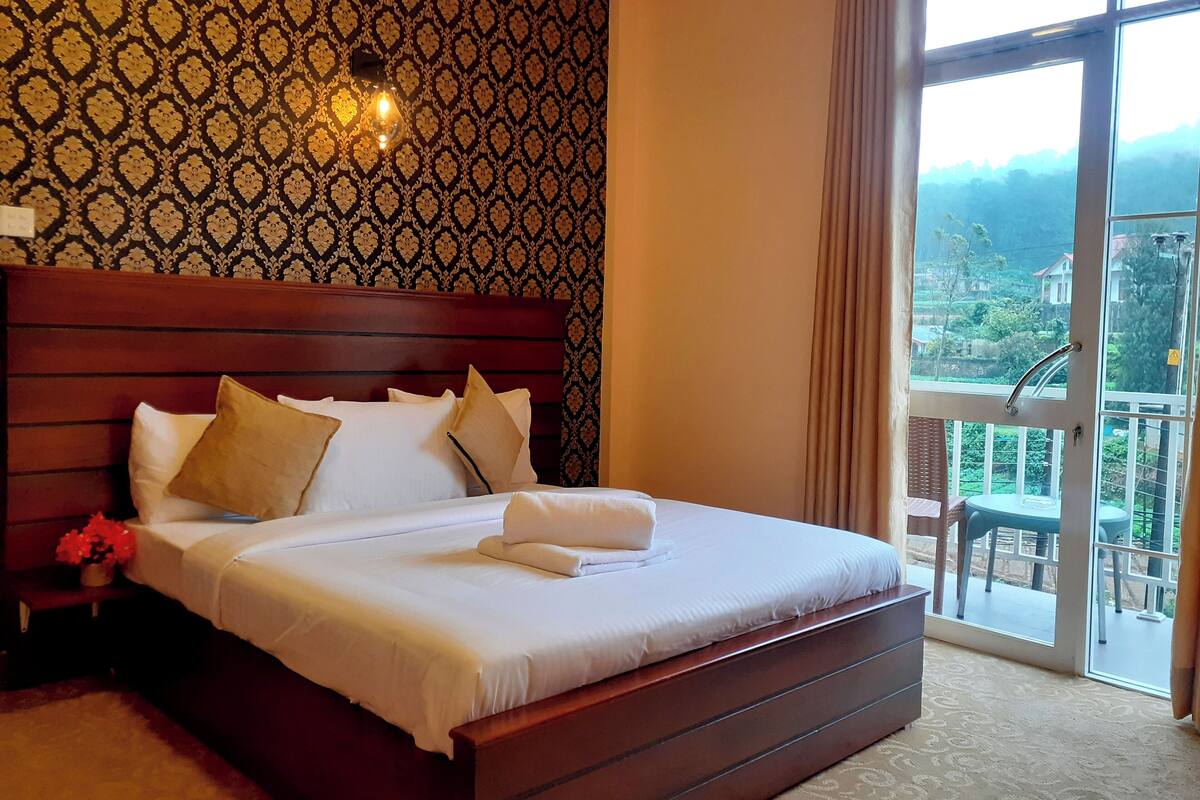 Ein Hotelzimmer mit einem großen Bett, einem Holz-Kopfschild, einem Nachttisch mit Lampe und Blick ins Freie durch ein großes Fenster.