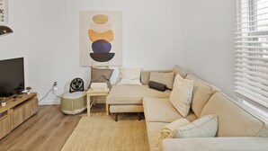 Un salon moderne avec un canapé beige, une petite table d’appoint, un ventilateur et un tableau accroché au mur.