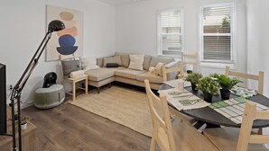 Un salon moderne comprenant un canapé d’angle, une table à manger avec des chaises, un ventilateur de plafond et une grande fenêtre avec des stores.