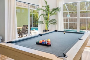 A pool table with a set of billiard balls, a potted plant, and a view of a swimming pool through a glass door. - Private heated  pool house with mini golf (Kissimmee)