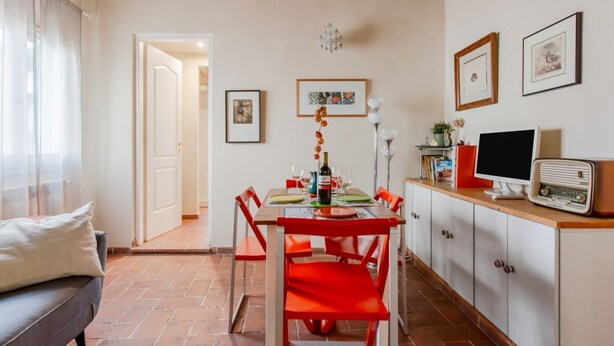 A dining area with a table set for two, red chairs, a white cabinet with a television, and framed pictures on the wall.
