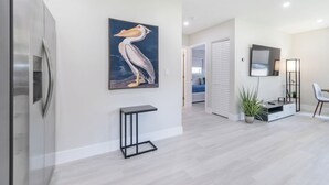 A modern kitchen with a stainless steel refrigerator, a painting of a pelican, a small black table, and a hallway leading to a bedroom. - TWO Casitas | Victoria Park | 30+ Night Stays Only (Fort Lauderdale)