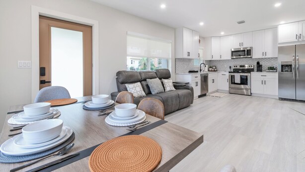 A modern kitchen with white cabinets, stainless steel appliances, and a dining area with a wooden table set for four. - TWO Casitas | Victoria Park | 30+ Night Stays Only (Fort Lauderdale)