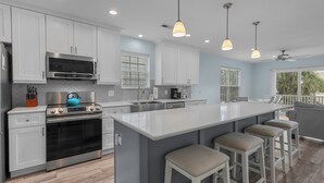 A modern kitchen with white cabinets, stainless steel appliances, and a large island with bar stools. - Island Girl-Beautiful 4BD Ocean View-Private Pool-Pet Friendly (Surfside Beach)