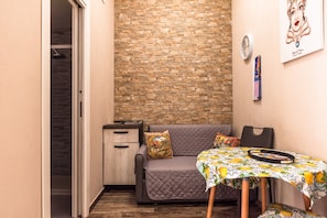 Une pièce chaleureuse avec un mur en pierre, un canapé, une table recouverte d’une nappe à motifs de fruits et une horloge au mur.