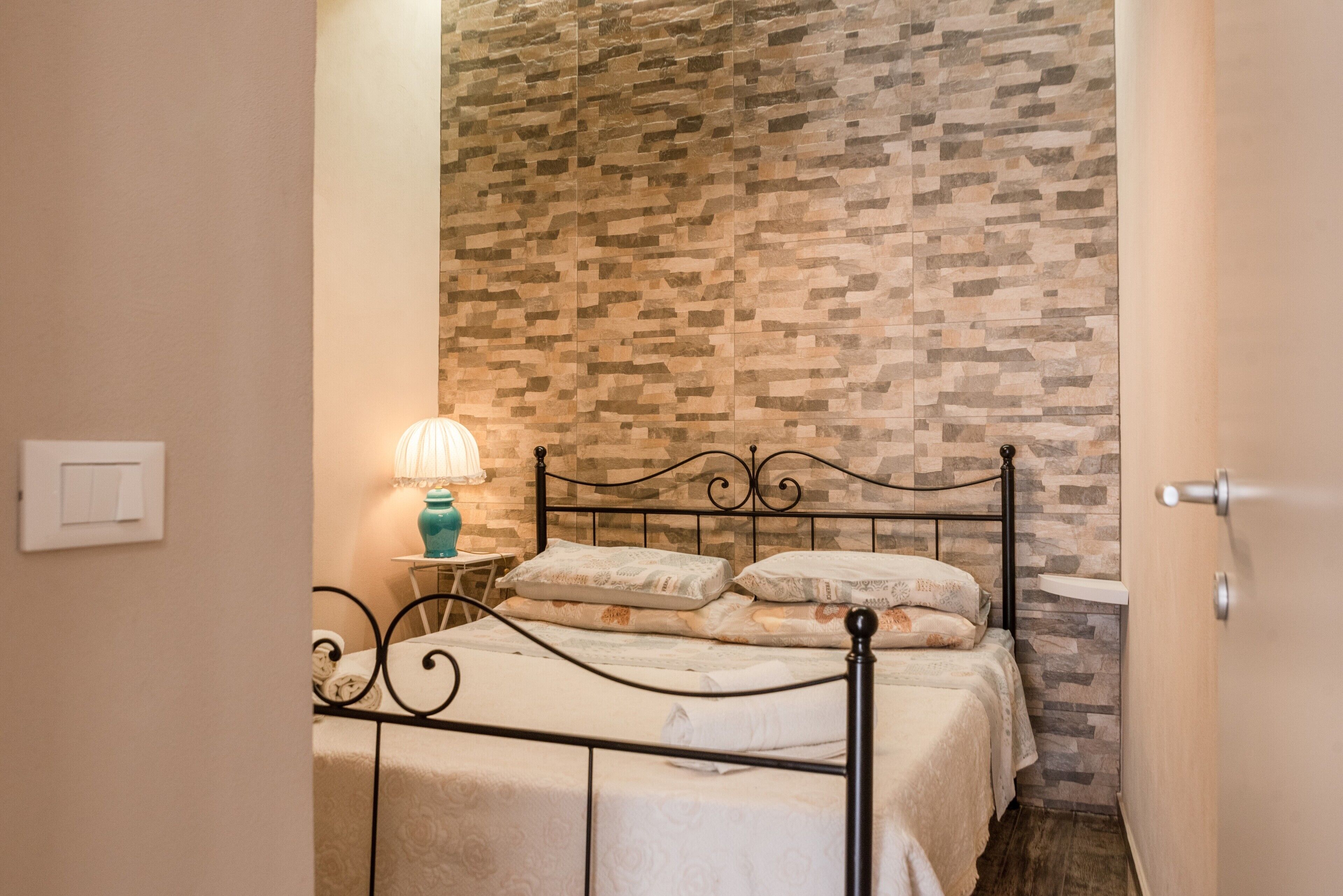 Quarto com parede em padrão de pedra, cama com estrutura de metal, luminária de cabeceira e uma pequena mesa.