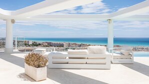 Una terraza en la azotea con mobiliario blanco, una planta en maceta y vistas al mar y al paisaje urbano.