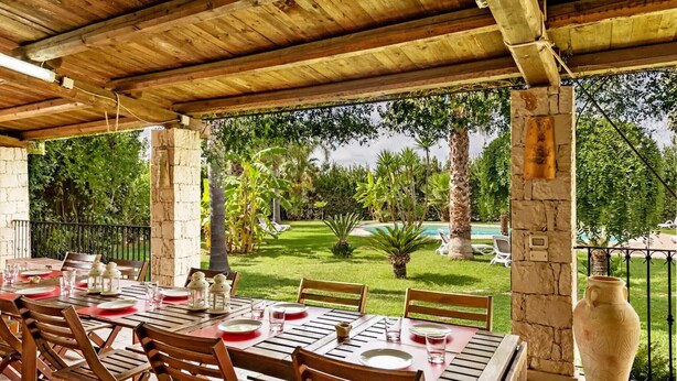 An outdoor dining area with stone pillars, wooden tables, and chairs, overlooking a well-maintained lawn and a swimming pool.