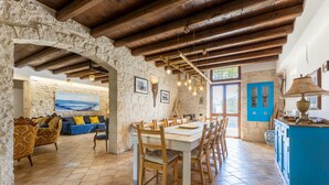 A rustic interior with stone walls, wooden beams, a dining area with a table and chairs, a sofa with cushions, and a kitchen area with blue cabinets.