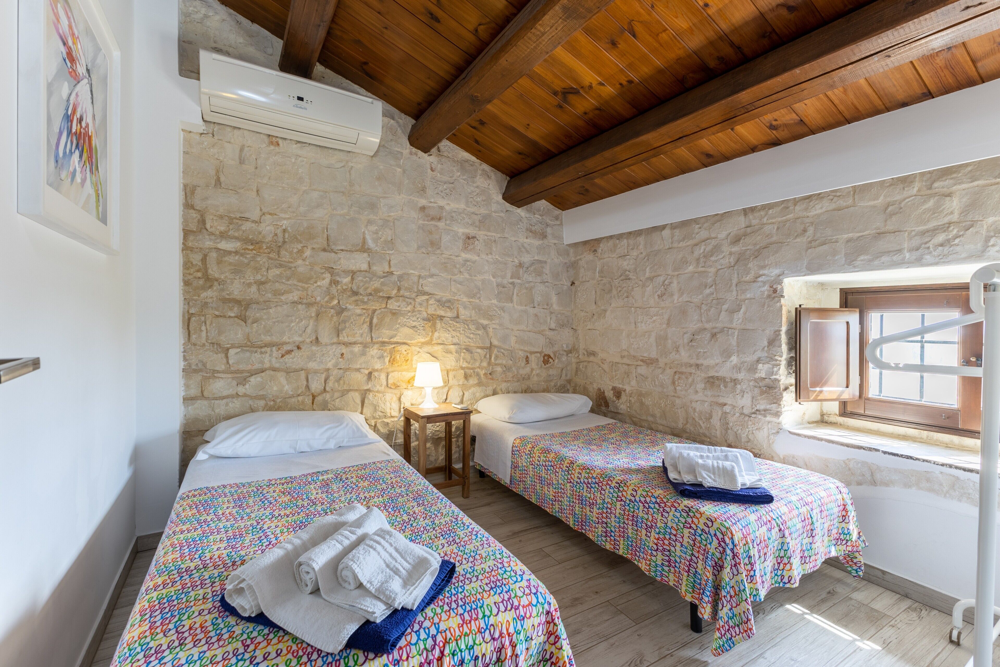 Ein Zimmer mit zwei Betten, einer Steinwand, Holzbalken, einem Fenster und einer Lampe.