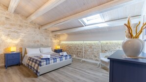 Ein Schlafzimmer mit Steinwand, Holzbalkendecke, Oberlicht, einem Bett mit karierter Decke, Nachttischlampen und einer dekorativen Vase.