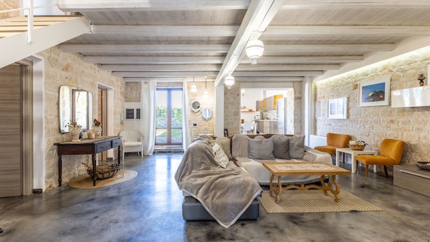 Ein geräumiger Wohnbereich mit Sofa, Couchtisch und Sesseln, im Hintergrund eine Küche und eine Steinwand.