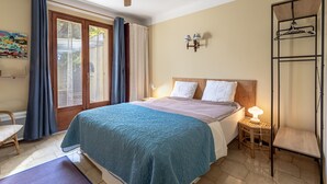 Een slaapkamer met een bed, een plafondventilator, een raam met gordijnen, een nachtkastje met een lamp en een kapstok aan de muur.