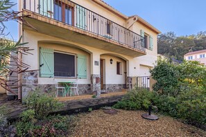 A two-story house with a balcony, a stone facade, and a garden area with plants and a table.