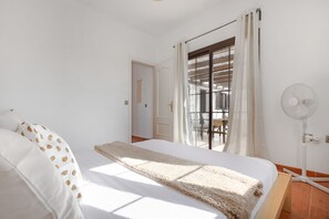 Un dormitorio con cama, ventana con cortinas, ventilador de pie y puerta que da a otra habitación.