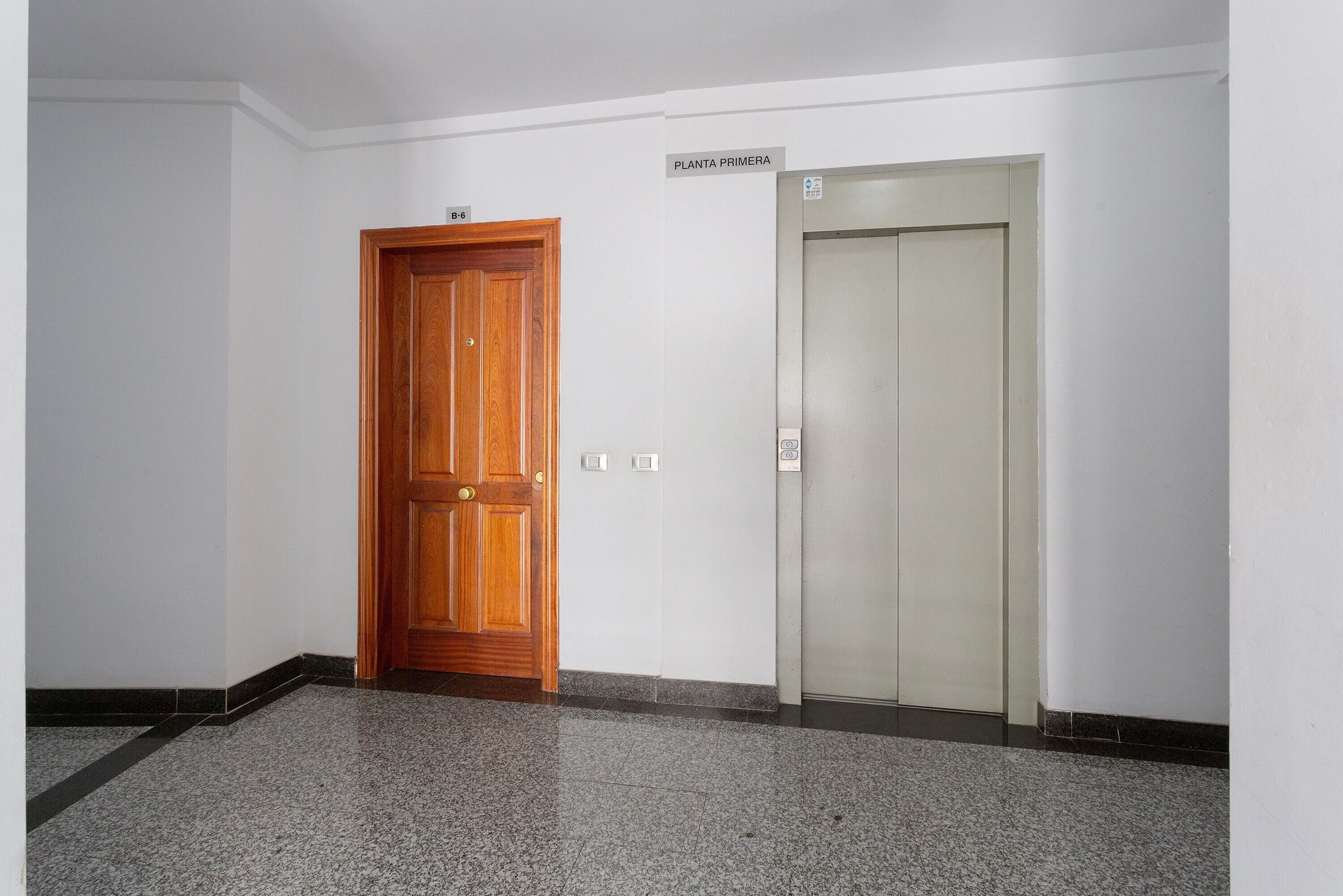 Un pasillo con una puerta de madera con la etiqueta 'B-6' y un ascensor con un cartel que dice 'PLANTA PRIMERA'.