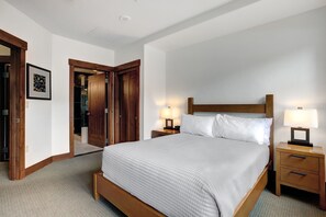 Habitación de hotel ordenada con una cama grande, cabecera de madera, dos mesitas de noche con lámparas, un cuadro enmarcado en la pared y una puerta de madera que da a otra habitación.