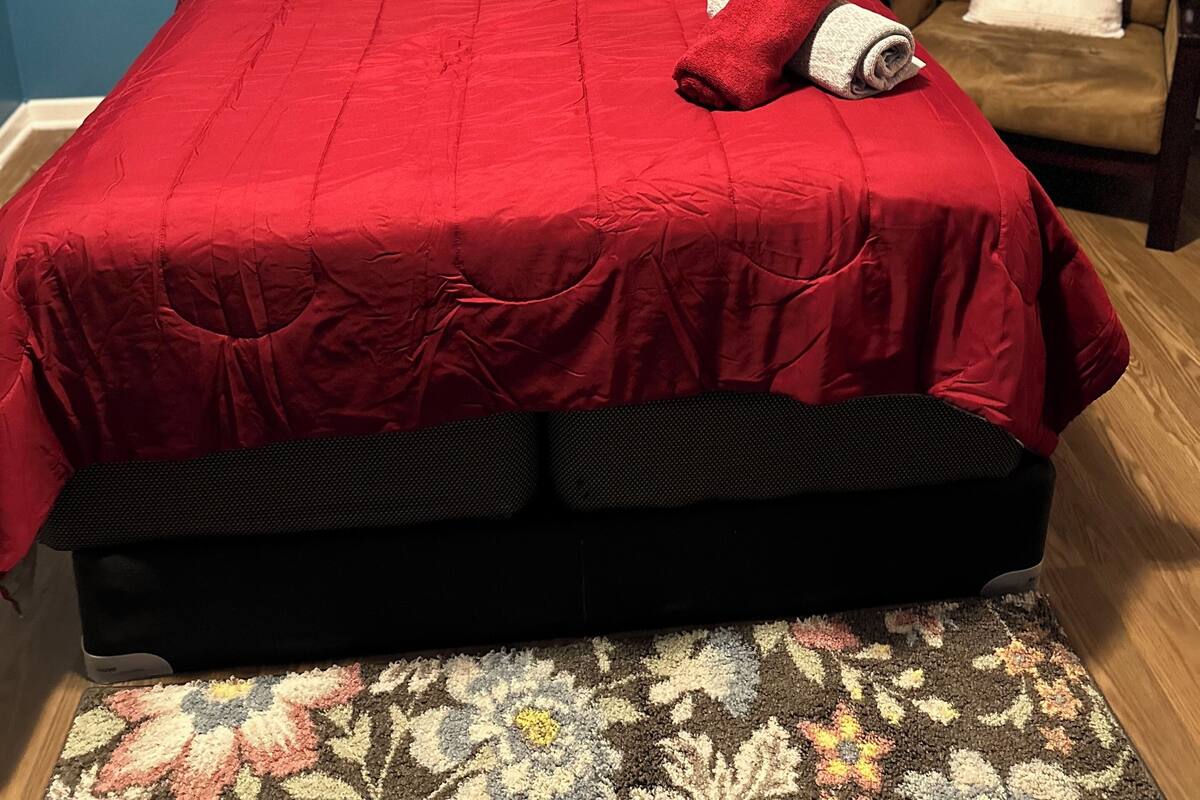 A bed with a red bedspread and pillows, a rolled-up towel, a chair with a cushion, and a picture on the wall.